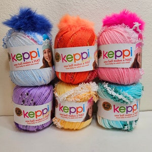 Peut inclure: Six pelotes de laine de différentes couleurs, dont bleu, rose, orange, jaune, violet et vert. Chaque pelote est surmontée d'un pompon en fourrure et porte une étiquette indiquant "keppi one ball makes a HAT from baby to adult size small."