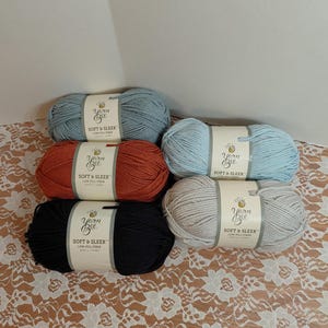 Puede incluir: Seis madejas de hilo de fibra baja Yarn Bee Soft & Sleek en varios colores: azul claro, óxido, negro, azul claro, gris y azul oscuro. Cada madeja tiene una etiqueta blanca con texto negro.