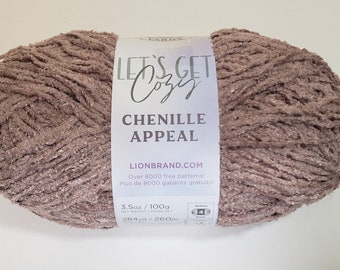 Può includere: Un gomitolo di filato di ciniglia in un colore malva tenue. L'etichetta recita "Let's Get Cozy Chenille Appeal" e "Lionbrand.com". Il filato pesa 100 g ed è lungo 260 m.