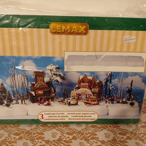 May include: A Lemax landscape accents set in a box. The box features a winter village scene with buildings, trees, and figures. The packaging includes the Lemax logo and the words "Landscape Accents" in multiple languages.