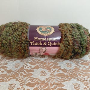 May include: A skein of Lion Brand Homespun Thick & Quick yarn in a variegated blend of olive green, brown, and tan. The center paper label features the brand logo, the text "Homespun Thick & Quick," and a photograph of a person holding yarn.