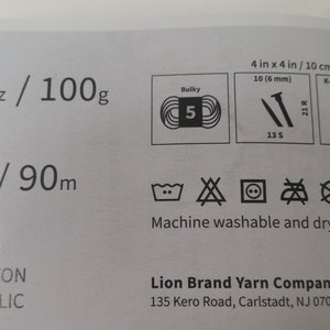 May include: A white label with black text and symbols. The label includes information about yarn weight, fiber content, and care instructions. The text reads "3.5 oz / 100g", "98yd / 90m", "69% Cotton", "31% Acrylic", "Machine washable and dryable", "Lion Brand Yarn Company", "135 Kero Road, Carlstadt, NJ 07072".