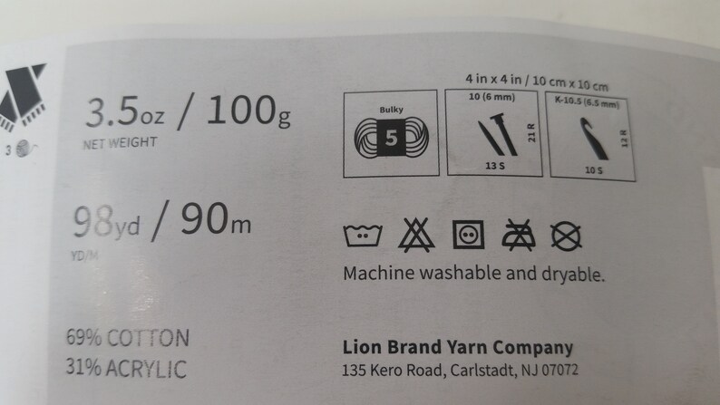 May include: A white label with black text and symbols. The label includes information about yarn weight, fiber content, and care instructions. The text reads "3.5 oz / 100g", "98yd / 90m", "69% Cotton", "31% Acrylic", "Machine washable and dryable", "Lion Brand Yarn Company", "135 Kero Road, Carlstadt, NJ 07072".