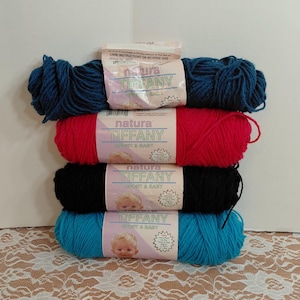 Könnte beinhalten: Vier Knäuel Natura Tiffany-Garn in den Farben Türkis, Rot, Schwarz und Marineblau. Jedes Knäuel ist mit dem Markennamen und einem Babybild versehen. Das Garn ist vertikal gestapelt.