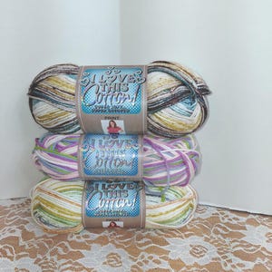 Puede incluir: Tres madejas de hilo estampado "I Love This Cotton!". La madeja superior presenta tonos marrones, beige y azules. La madeja del medio tiene tonos rosas, morados y verdes. La madeja inferior muestra rayas amarillas, verdes y blancas.