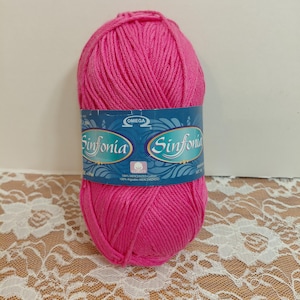 May include: A skein of bright pink yarn wrapped with a blue label featuring the Omega logo and the text Sinfonia. The label specifies 100% mercerized cotton. The yarn is wound into an oval shape with visible strands of the vibrant pink fiber.