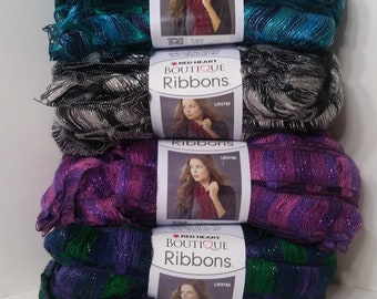 May include: Four skeins of Red Heart Boutique Ribbons yarn in different colours: teal, silver, purple, and green. Each skein has a label with a woman wearing a scarf made from the yarn.
