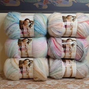 Puede incluir: Seis ovillos de hilo multicolor en tonos de rosa, azul y amarillo. El hilo está etiquetado como "scarfie" y presenta a una mujer con una prenda de punto de ganchillo.