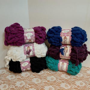 Könnte beinhalten: Eine Sammlung farbenfroher Garnknäuel in Lila, Weiß, Schwarz, Marineblau, Türkis und Burgunderrot. Jedes Knäuel ist mit einem Etikett mit einer Frau versehen. Das Garn wirkt weich und dick, geeignet für verschiedene Strick- oder Häkelprojekte.