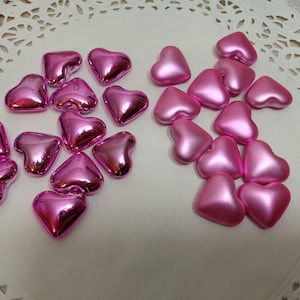 Könnte beinhalten: Eine Sammlung von rosa herzförmigen Perlen mit metallischem Glanz. Die Perlen sind auf einer weißen Oberfläche mit einem spitzenartigen Muster verstreut. Die Herzen variieren leicht in Form und Größe.