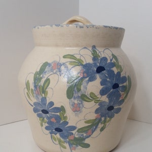 以下が含まれることがあります： クリーム色のセラミックジャー、蓋とハンドル付き。ジャーは、青い花、緑の葉、ピンクのアクセントのリースで飾られています。ジャーはわずかにテクスチャ加工された表面とスカラップの縁を持っています。