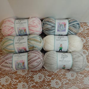 以下が含まれることがあります： さまざまな色のBaby Bee Sweet Delight Printsの糸が6玉。糸は花柄の表面に配置されています。各玉にはブランド名と赤ちゃんの写真が記載されたラベルが付いています。