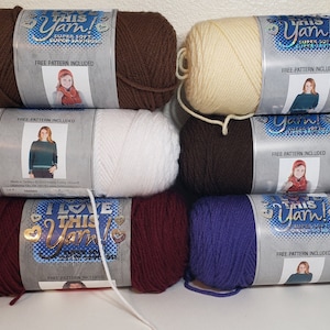 Peut inclure: Une pile de huit pelotes de laine de différentes couleurs, dont le brun, le blanc, le bordeaux et le violet. Chaque pelote porte une étiquette indiquant "I Love This Yarn!" avec un modèle gratuit inclus.