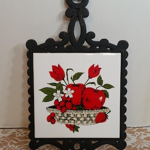May include: Black cast iron trivet with a white tile insert featuring a fruit basket design. The tile shows red apples, grapes, strawberries, and tulips. The trivet has a decorative, scalloped edge and a handle.