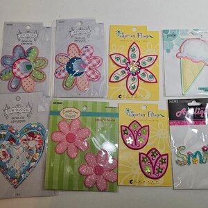 May include: A collection of iron-on appliques featuring floral and other designs, including a heart, an ice cream cone, and the word "Smile". The appliques use colourful fabrics, sequins, and the text "Sew Ology", "Spring Fling", and "Creations Grace".