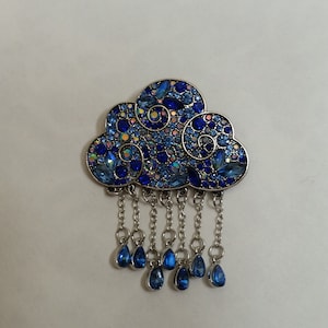 May include: A silver-toned brooch shaped like a cloud with blue and iridescent rhinestones. The cloud has a dangling chain with blue teardrop-shaped crystals.