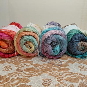 May include: Four skeins of colorful yarn in shades of pink, orange, teal, and blue. Each skein is rolled into a tight coil, with a label visible on top. The yarn is arranged on a lace tablecloth.
