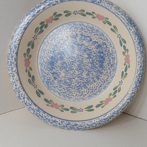 Może przedstawiać: Okrągły, ceramiczny talerz z niebieskim, nakrapianym środkiem i kremowym obramowaniem. Obręcz ma niebieski, nakrapiany wzór. Różowe serca i zielone liście zdobią kremowe obramowanie. Dekoracyjny element do kuchni.