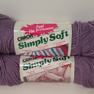 Può includere: Due gomitoli di filato Caron Simply Soft di colore lilla chiaro. Il filato è confezionato in un sacchetto di plastica con un'etichetta che recita "Senti la differenza Caron Simply Soft" e elenca le caratteristiche del filato: "Morbidezza superiore", "Colori distintivi" e "Qualità impareggiabile".