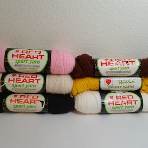 Puede incluir: Una pila de ocho ovillos de hilo Red Heart sport en varios colores, incluyendo rosa, blanco, marrón, amarillo y negro. El hilo es lavable a máquina y secable en secadora. La etiqueta dice "Red Heart Sport Yarn" y "Wintuk Sport Yarn".