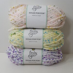 Puede incluir: Tres ovillos de hilo Baby Bee Sugar Pom Pom en rosa, amarillo y morado con motas blancas. Cada ovillo tiene una etiqueta con el nombre de la marca y el tipo de hilo.