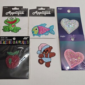 May include: Selection of iron-on appliques including a green frog, a fish, a teddy bear, a red apple, and two sequined hearts. These colourful appliques are designed for wearable art and craft projects.