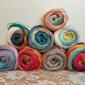 Op de afbeelding: Een verzameling van negen bollen garen gerangschikt in een piramidestapel. Elke bol heeft een meerkleurig verloopmotief, inclusief tinten roze, blauw, turquoise, geel, oranje, paars en grijs, met een gladde, gedraaide vezelstructuur door het geheel.