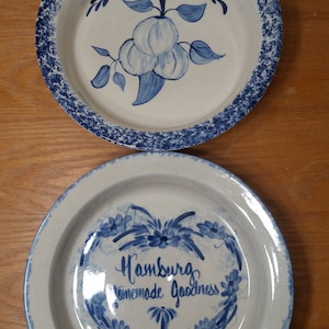 Puede incluir: Dos platos para tartas de cerámica azul y blanca. Un plato tiene un diseño de melocotón azul y blanco con el texto "Peach Pie". El otro plato tiene un diseño floral azul y blanco con el texto "Hamburg Homemade Goodness".