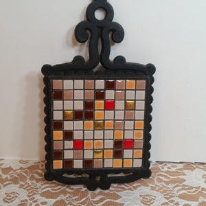 Puede incluir: Un salvamanteles decorativo con marco y asa de hierro fundido negro. El centro presenta un mosaico de pequeñas baldosas cuadradas en tonos marrón, beige, rojo y dorado. El salvamanteles está diseñado para proteger superficies.