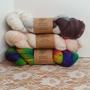 May include: Three skeins of hand-dyed yarn in a stack. The top skein is white with black speckles, the middle is a light peach, and the bottom is a vibrant rainbow of colors. Each skein has a brown paper label that reads "Yarn Bee Authentic Hand-Dyed".