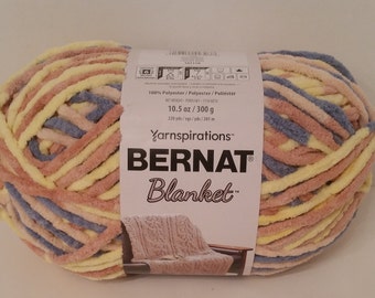 Puede incluir: Ovillo de lana Bernat Blanket en colores pastel. El hilo es una mezcla de amarillo, rosa y azul. La etiqueta indica 300 g. El hilo es 100% poliéster.