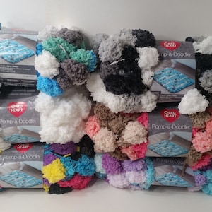 May include: A stack of yarn skeins in various colors, including white, gray, black, teal, pink, and purple. The yarn is labeled "Pomp-a-Doodle" and is made by Red Heart.