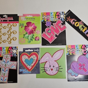 Könnte beinhalten: Eine Sammlung bunter Aufbügler, darunter goldene Wirbel, eine rosa Hibiskusblüte, das Wort "LOVE", ein Kreuz, ein Herz, ein Hase, ein Herz mit Flügeln und der Text "Go Girl".