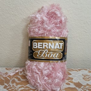 May include: A skein of light pink Bernat Boa yarn with a fuzzy, feathery texture. The black and gold label features the brand name Bernat and the product name Boa in white text. The yarn has a shiny, ribbon-like appearance throughout the ball.