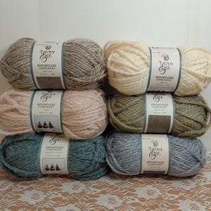 May include: Six skeins of Yarn Bee Effortless Super Bulky yarn stacked in two columns. The colors include beige, cream, light pink, olive green, teal, and grey. Each skein features a white paper label with the brand name and product details.