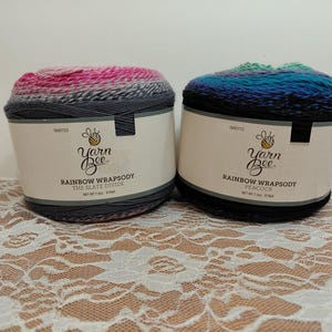 May include: Two skeins of Yarn Bee Rainbow Wrapsody yarn. The left skein, labeled The Slate Divide, features a gradient of pink, gray, and dark charcoal. The right skein, labeled Peacock, features a gradient of green, blue, and black.
