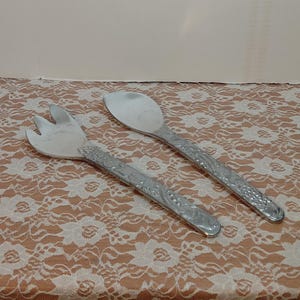 May include: A pair of silver salad servers. One server is a spoon, and the other is a fork. Both have ornate, textured handles with a floral design. The servers are displayed on a peach-colored lace tablecloth.