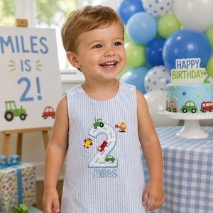 May include: A light blue and white seersucker sleeveless romper featuring an embroidered large number two with small colorful vehicles and the name Miles stitched below. The romper has two white buttons on each shoulder strap for closure.