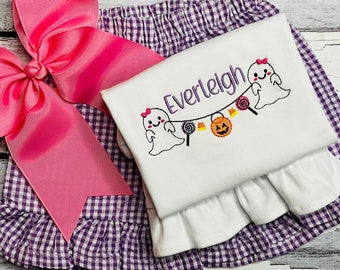 May include: A white shirt with a purple and white gingham skirt. The shirt has a ghost and candy corn garland with the name "Everleigh" embroidered on it. A pink bow is on top of the skirt.