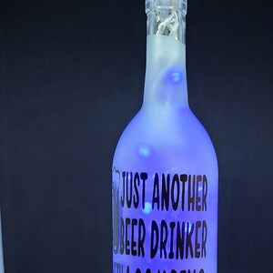 May include: A frosted glass bottle with a crystal stopper, illuminated with blue lights. The bottle has the text "JUST ANOTHER BEER DRINKER WITH A CAMPING PROBLEM" and a camper graphic.