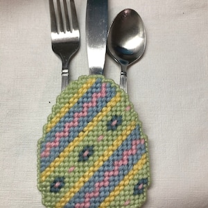 May include: A colorful Easter egg-shaped needlepoint design with a green, yellow, blue, and pink pattern. A fork, knife, and spoon are placed in the design.