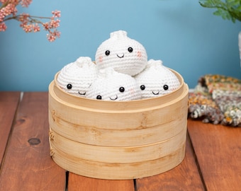 May include: Five white crocheted dumplings with smiling faces are arranged in a round bamboo steamer. The steamer is made of natural wood and has three tiers.