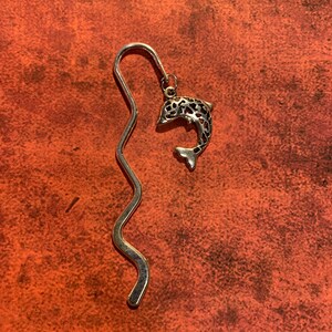May include: Silver bookmark with a dolphin design. The bookmark is shaped like a wave and has a delicate, intricate design.