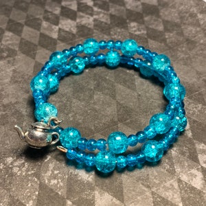 Puede incluir: Una pulsera envolvente con dos hilos de cuentas azules craqueladas. Se adjunta un pequeño dije de tetera plateada. La pulsera se envuelve alrededor de la muñeca. Las cuentas son de un vibrante color turquesa.