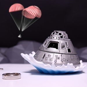 Può includere: Un portagioie a forma di capsula spaziale color argento è posto su una base bianca e blu, con due anelli e paracadute sullo sfondo. Il portagioie presenta dettagli ed è ambientato su uno sfondo scuro con formazioni rocciose grigie.