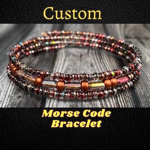 Peut inclure: Un bracelet multi-rangs composé de petites perles irisées dans des tons de brun, rouge et or. Le bracelet est enroulé autour d'une surface en bois et porte le texte "Custom Morse Code Bracelet".