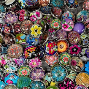 Puede incluir: Una colección de botones de presión coloridos con diversos diseños florales y geométricos. Los botones presentan un borde de metal plateado y una parte superior abovedada, mostrando colores vibrantes como rosa, amarillo, azul y naranja.