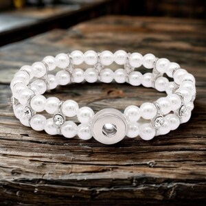 May include: A white pearl bracelet with three rows of faux pearls. The bracelet has a silver-colored snap closure and is accented with small, clear rhinestones. The bracelet is displayed on a wooden surface.