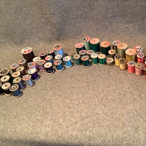 May include: A collection of vintage wooden spools of thread in various colors, including blue, green, pink, red, and white. The spools are arranged on a gray surface, showcasing a variety of thread types and hues, perfect for sewing and crafting projects.