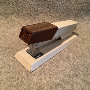 Può includere: Una graffatrice bianca e marrone con un manico in legno. La graffatrice ha una base in metallo e un estrattore di graffe argentato. La graffatrice è etichettata "Bates 221 Heavy Duty".
